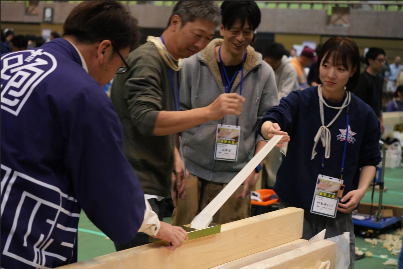 作業服姿の4人が、木材の表面を削る「削ろう会」のイベント会場で、手前の男性が鉋を使って削り出した極めて薄い木片を、笑顔の女性と中央の男性が驚いた表情で見つめる様子を写した写真