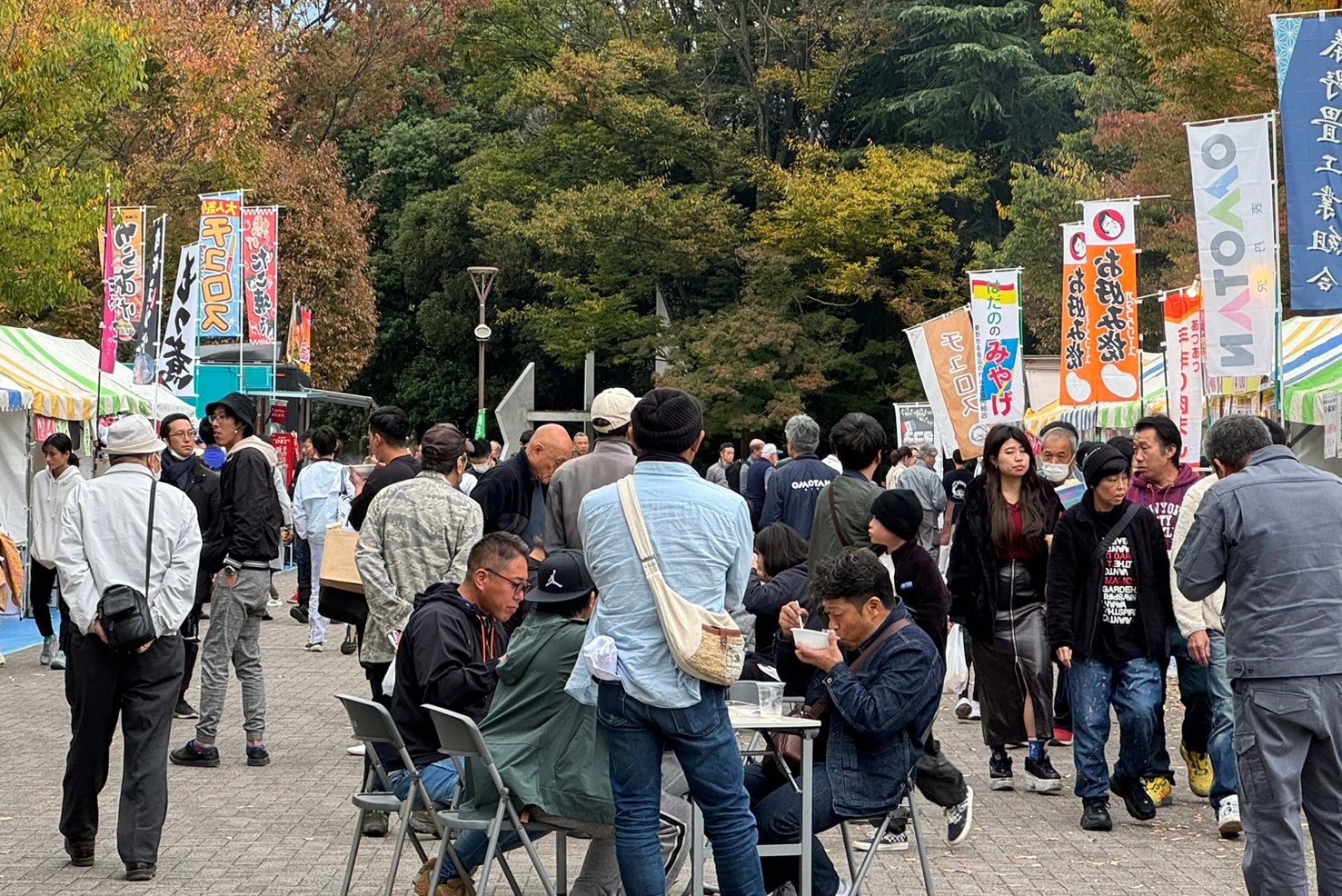 秋の木々に囲まれた屋外の広場で開催されている賑やかなイベント会場で、多くの来場者が屋台の前に立ち、テーブルで食事をして交流する様子の祭り風景の写真