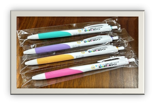 木目調テーブルに整然と並べられた透明パッケージ入りの四色(緑・紫・黄・ピンク)のグリップ部分を持つボールペンセットの文具写真