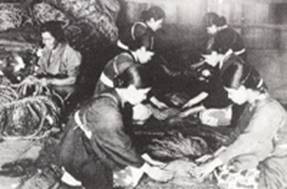 土間で藁のような素材を囲み座った和服姿の女性たちが、薄暗い室内で手作業に熱中している様子を捉えたモノクロ写真