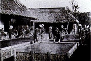 茅葺き屋根の建物の前で、数人の人々が囲まれた屋外の広い作業台を囲んで何らかの作業をしている様子を写したモノクロ写真