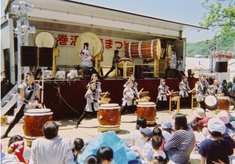 屋外のイベントで、トラックの荷台をステージに、揃いの衣装を着た人々が大小の和太鼓を力強く演奏し、多くの親子連れが鑑賞している写真