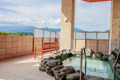 建物のバルコニーに設置された岩造りの湯船に湯が張られ、その横にベンチが置かれており、柵の向こうには雄大な山並みが広がっている露天風呂の写真