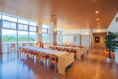 木目調の床と天井が印象的な広々としたラウンジで、左側の大きな窓からは緑豊かな山々が望め、長い木製テーブルと椅子が整然と並べられている明るい食堂の写真