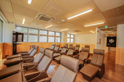 木目調の床と天井が特徴的な休憩室で、窓際にはテレビが置かれ、茶色いリクライニングチェアが並び、奥には「ゆ」の暖簾が見える無料休憩室の写真