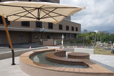 屋外の広場に、大きなベージュのパラソルが立ち、木目調のベンチと一体になった曲線を描く足湯が設置されている写真