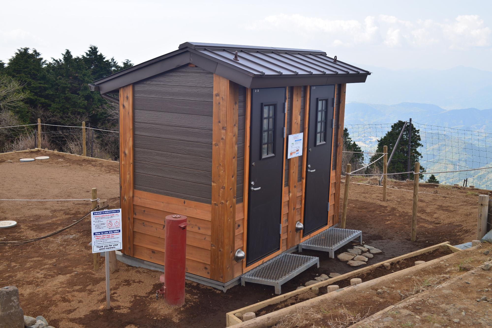 山の上に、木目調の壁と2つの黒いドアを持つ三ノ塔の公衆トイレの外観写真