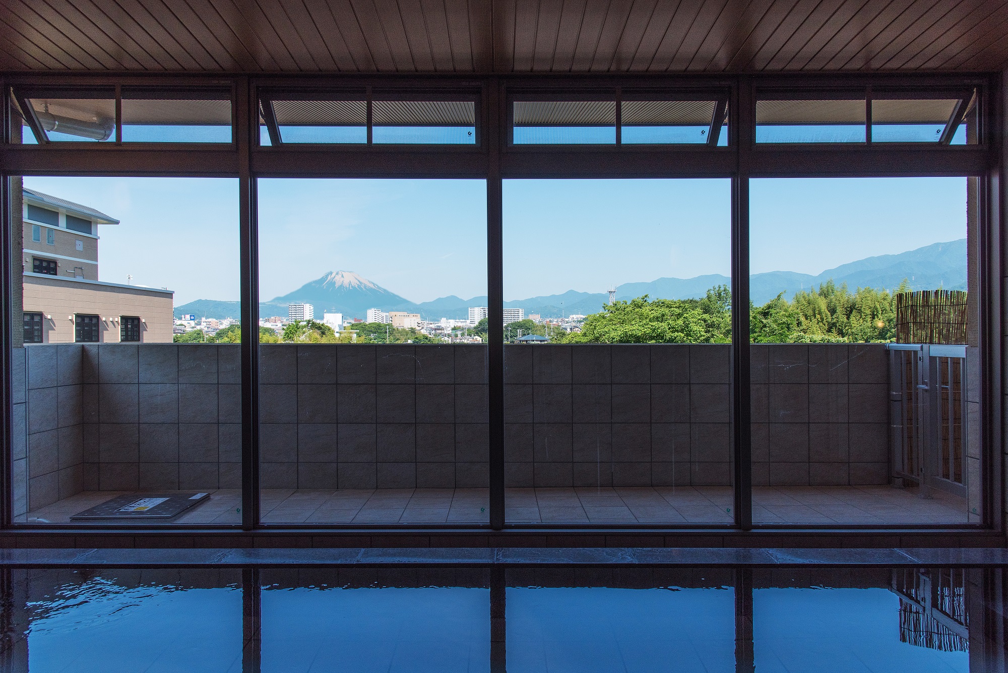 浴室内から見た大きな窓には手前の水面が映り込み、窓の向こうにはバルコニーと、雪が積もる富士山、街並み、緑豊かな山々が晴れた空の下に広がる浴場からの眺望の写真