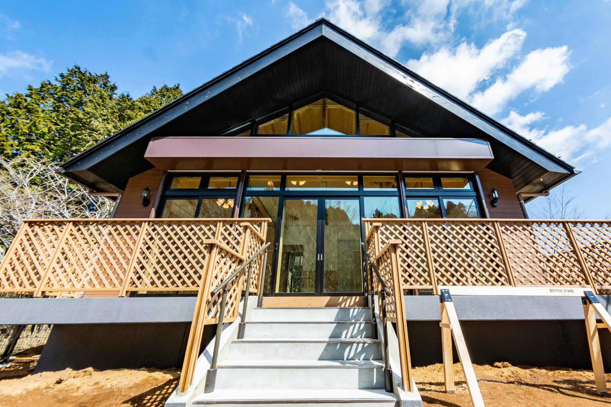 青空と雲が広がる晴れた日に、ウッドデッキを備えたモダンな山荘風のヤビツ峠レストハウスを、正面の階段下から見上げるように撮影した写真