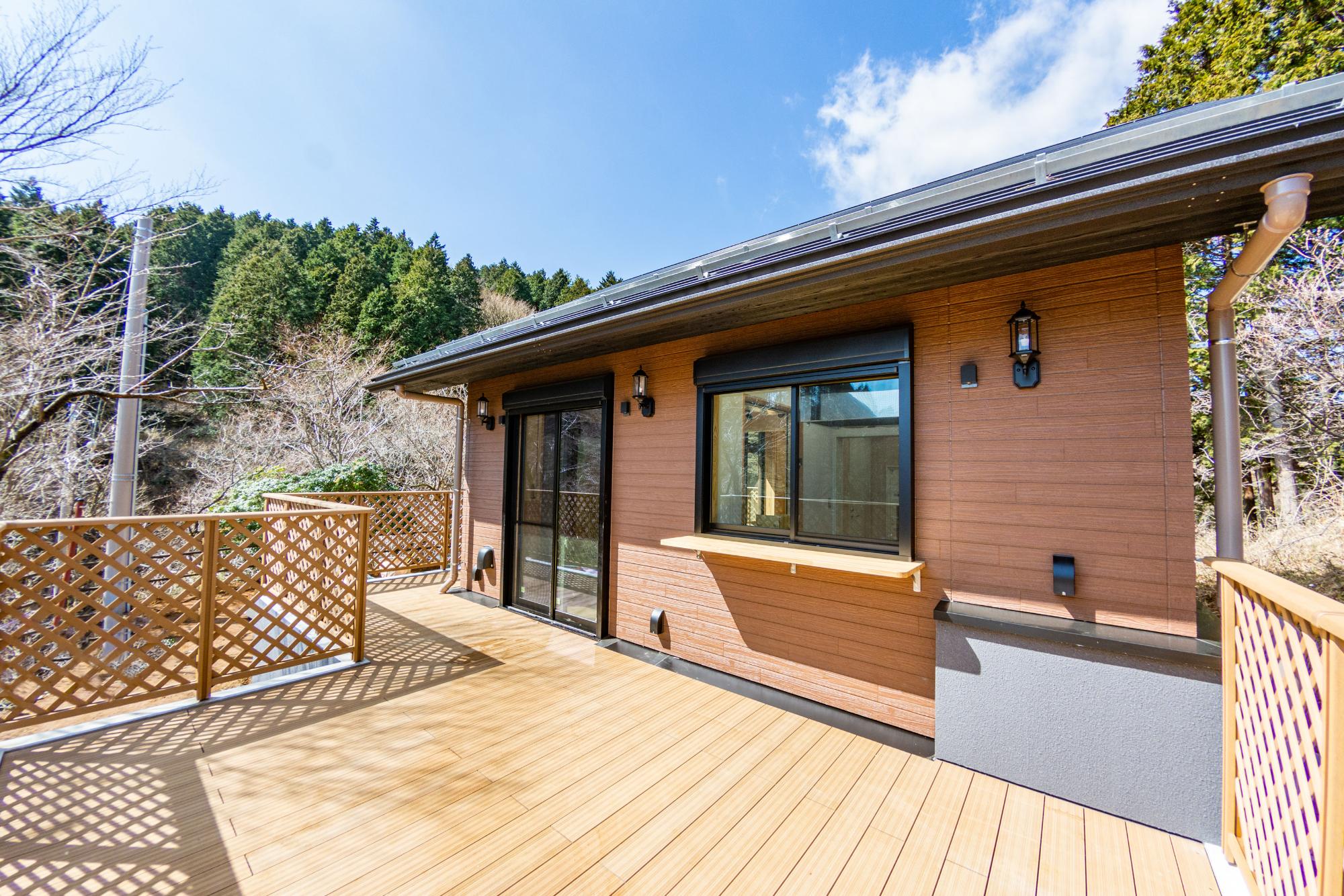 晴れた日に、森を背景にしたヤビツ峠レストハウスの広々としたウッドデッキと、窓やドアが設置された茶色い壁を横から撮影した構図の写真