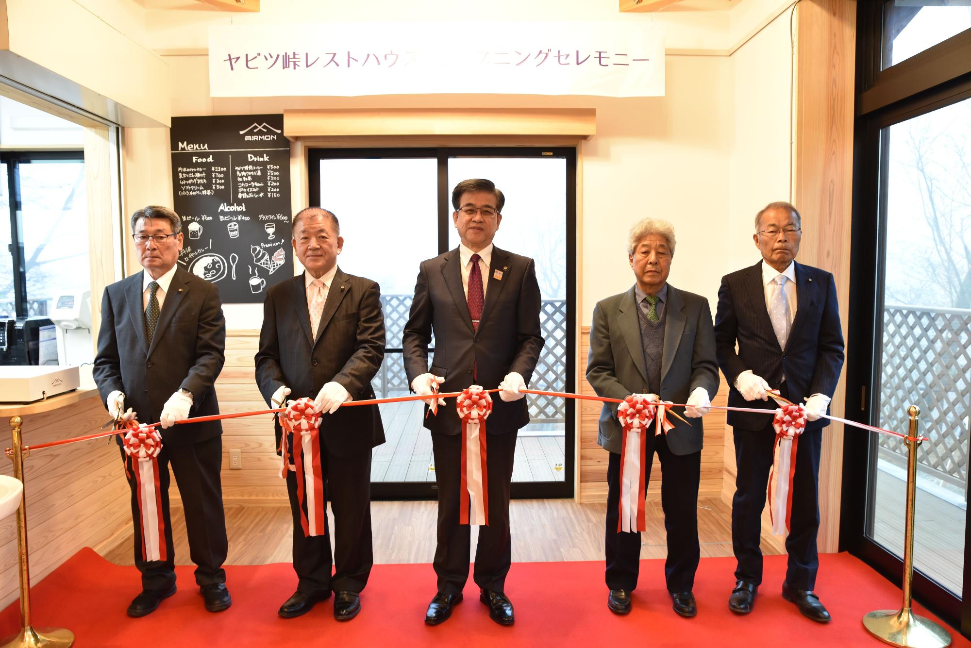 「ヤビツ峠レストハウス」のオープニングセレモニーで、市長を中心にスーツを着用した5人の男性が赤い絨毯の上で、これから行われるテープカットのために白い手袋をはめて紅白の大きなリボンの付いたテープを手にしている写真