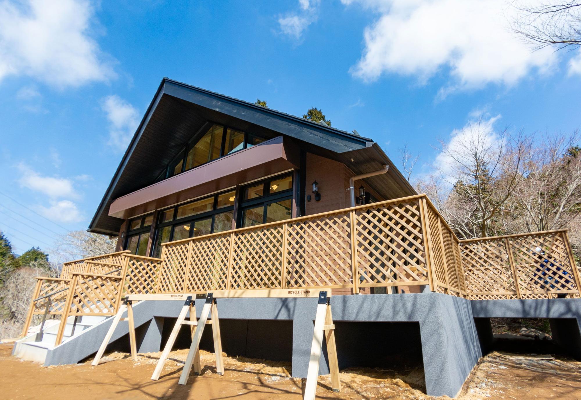 青空と白い雲が広がる晴れた日に、ウッドデッキが備え付けられたモダンな山荘風のヤビツ峠レストハウスを、低い位置から見上げた写真