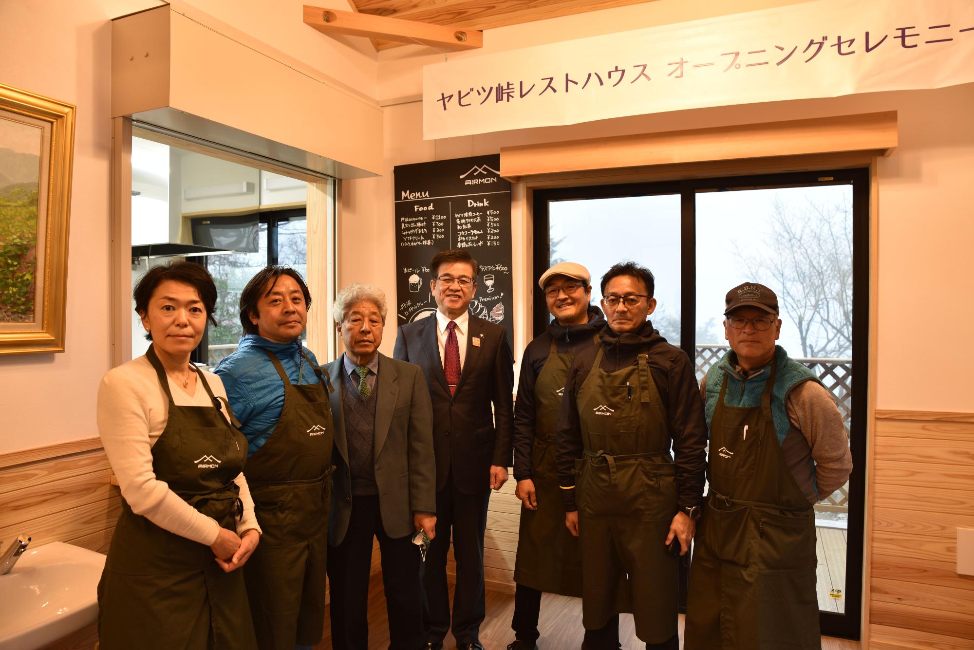 ヤビツ峠レストハウスのオープニングセレモニーで、関係者と思われるスーツ姿の男性やエプロンを着用した男女7人が、店内の黒板メニューの前で並んで記念撮影に応じている様子の写真
