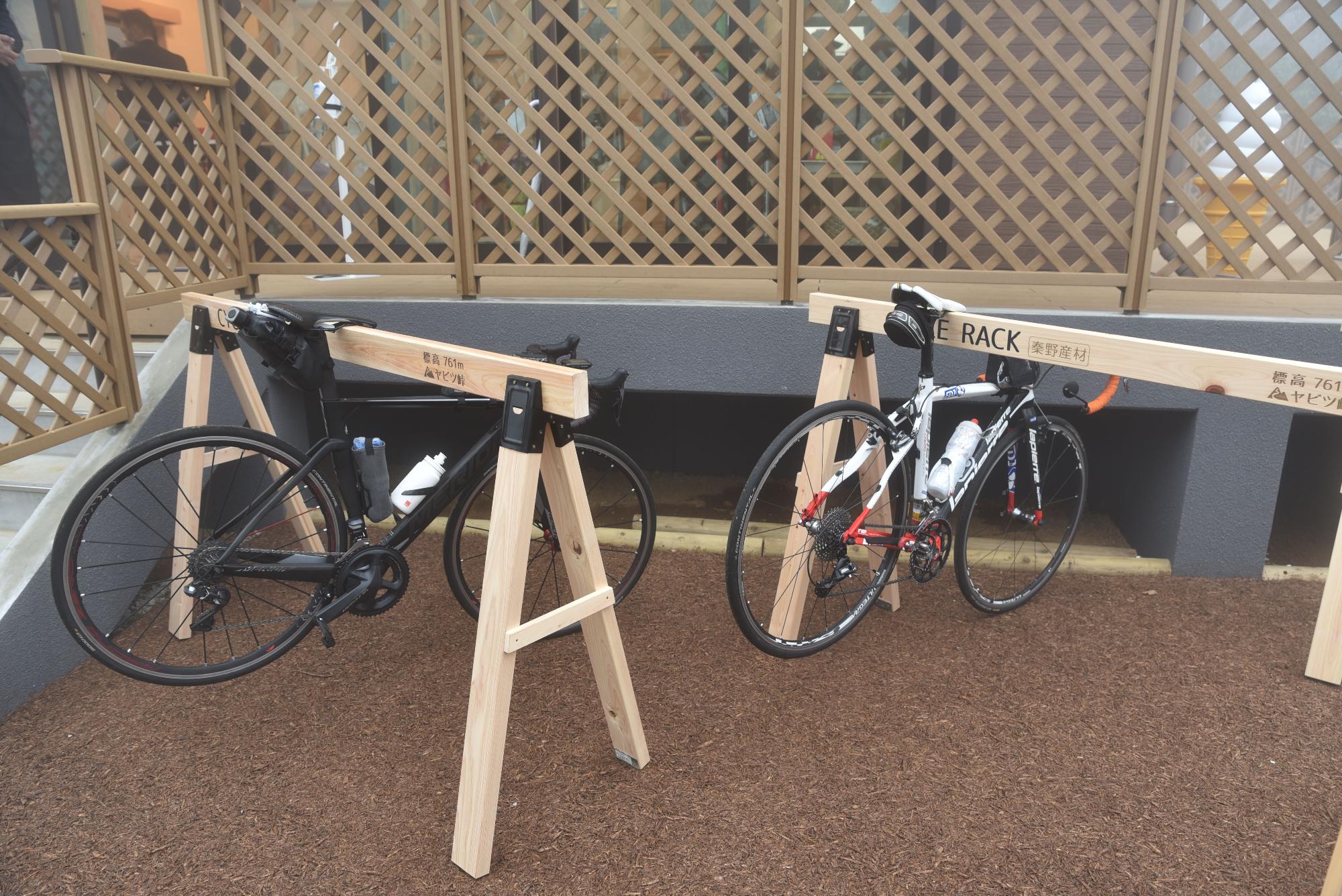 ウッドチップが敷かれた駐輪スペースに、秦野産材で作られ「ヤビツ峠」と記された木製サイクルラックに、黒と白のロードバイク2台が駐輪されている写真