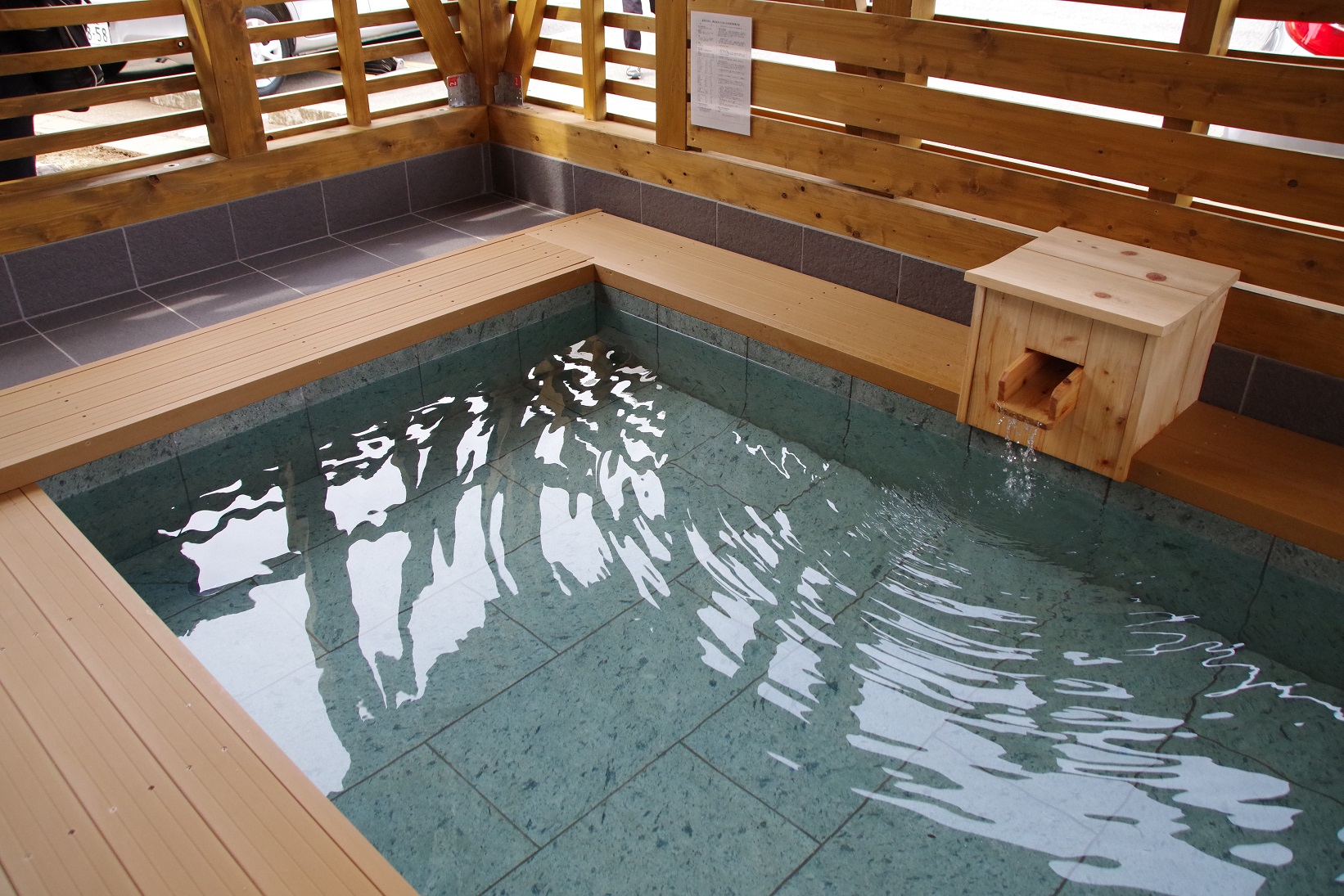 木製の柵で囲まれた半屋外の空間に水色のタイル張りの四角い湯船が設置され、縁には木製デッキがあり、右奥の木箱型湯口から注がれた湯で水面が波立っている足湯の写真