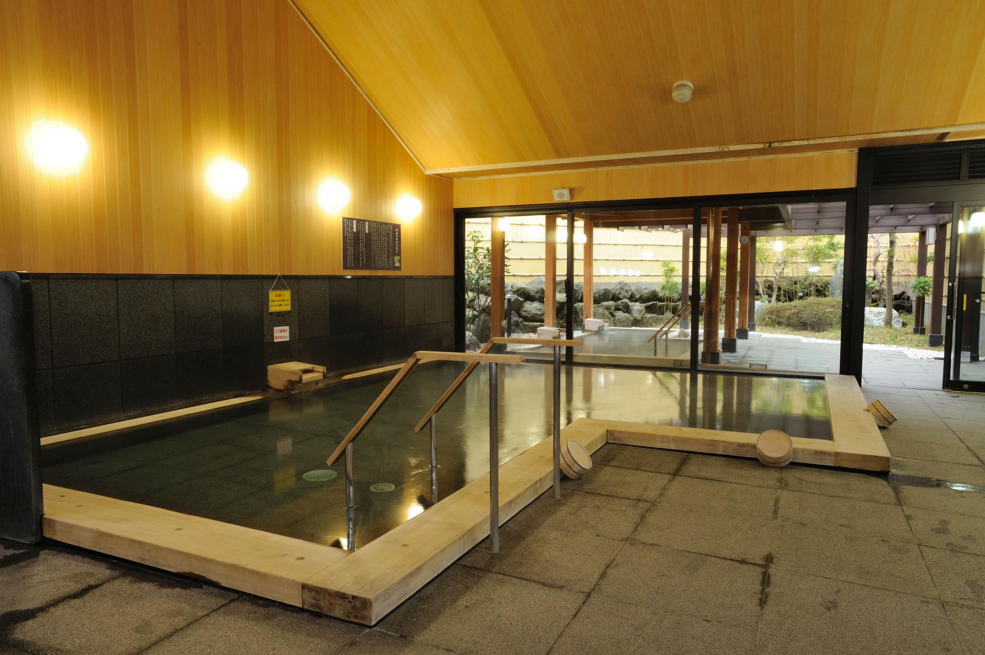 木の温もりある壁と天井に囲まれた室内浴場で、湯船の先にガラス越しの庭園と露天風呂が見える落ち着いた雰囲気の里湯の写真