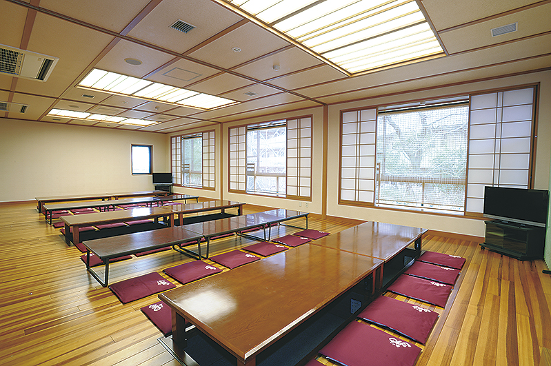 和風の広々とした宴会場で、明るい木目の床の上に低い木製の座卓とエンジ色の座布団が数列にわたって整然と並べられ、障子のある窓と格子状の天井照明が特徴的な貸切休憩室の写真
