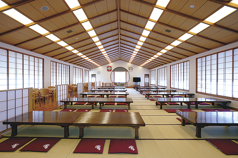 畳が敷かれた広々とした和風の宴会場で、低い座卓とエンジ色の座布団が数列に並べられ、両側には障子窓が並び、奥には丸窓とソファスペースが設けられている大広間の写真