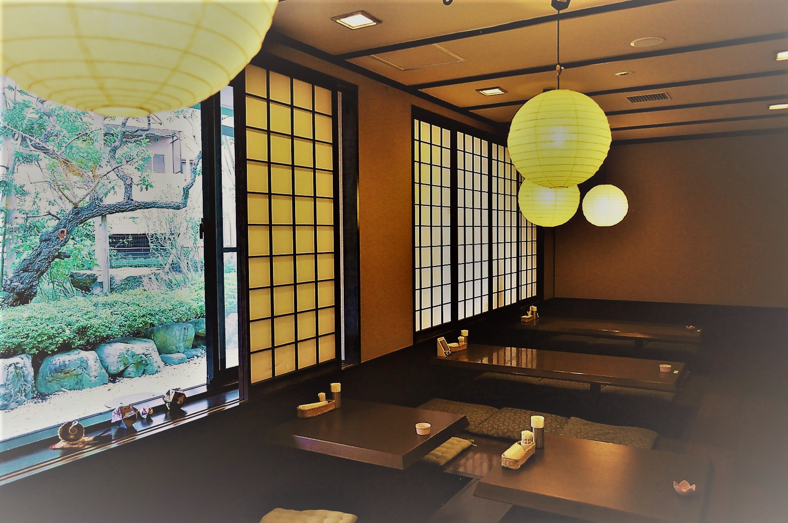 落ち着いた照明の和室に低い座卓と座布団が並び、開いた窓からは日本庭園が望め、天井からは複数の丸い紙の照明が吊り下げられている趣のある鶴寿庵の写真