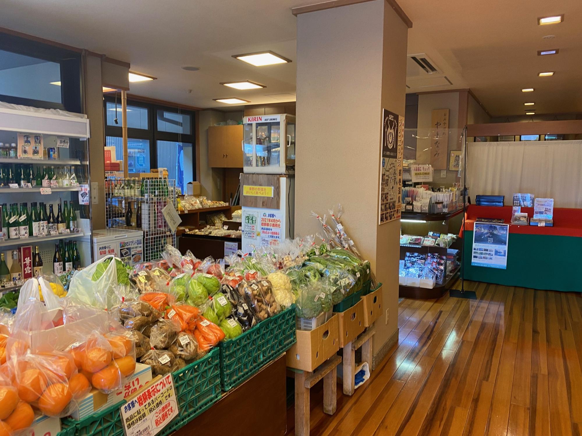 手前の緑のカゴにはみかんや人参、葉物野菜などの農産物が並び、奥にはお酒の棚や別の商品コーナーが見える店内の様子の写真