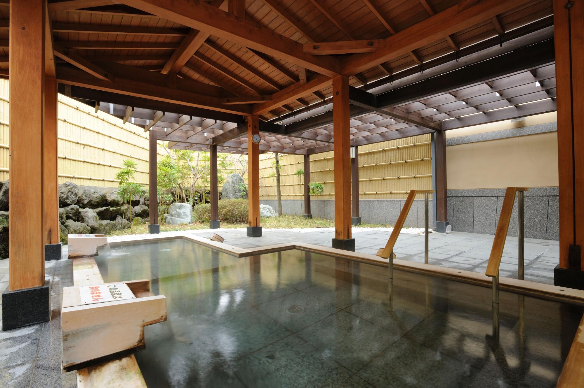 太い木の柱と屋根が印象的な和風の半露天風呂で、石造りの広い湯船に湯がたたえられ、奥には竹垣に囲まれた小さな庭が設えられている里湯の露天風呂の写真