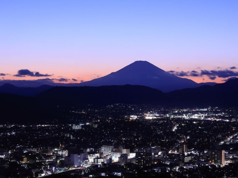 青とオレンジ色のグラデーションの空を背景に、雄大な富士山のシルエットが浮かび上がり、手前には無数の光が灯る市街地の夜景が広がっている写真