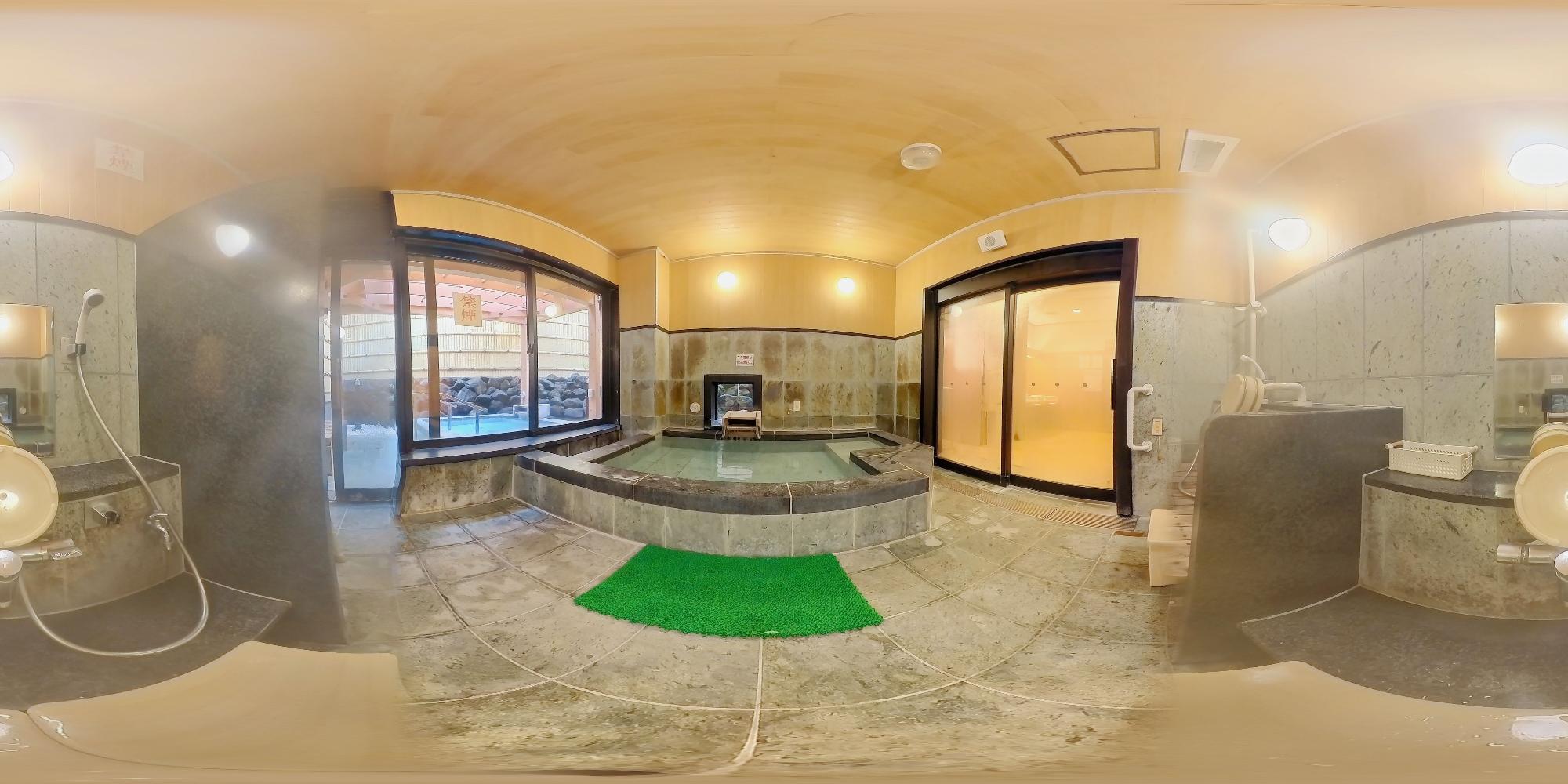 木の天井と石造りの壁に囲まれた小さな浴室で、中央の浴槽から窓越しに露天風呂が見える全方位視点で鶴の湯内湯を撮影した写真