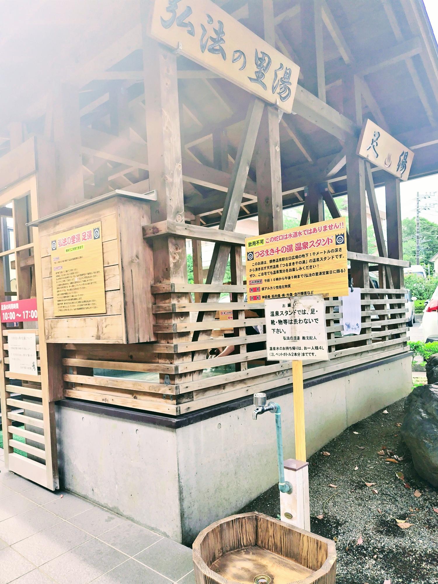 「弘法の里湯」と書かれた看板があり、利用案内の看板表示があり、手前には湯を汲むための蛇口と木の桶が置かれている温泉スタンドの写真