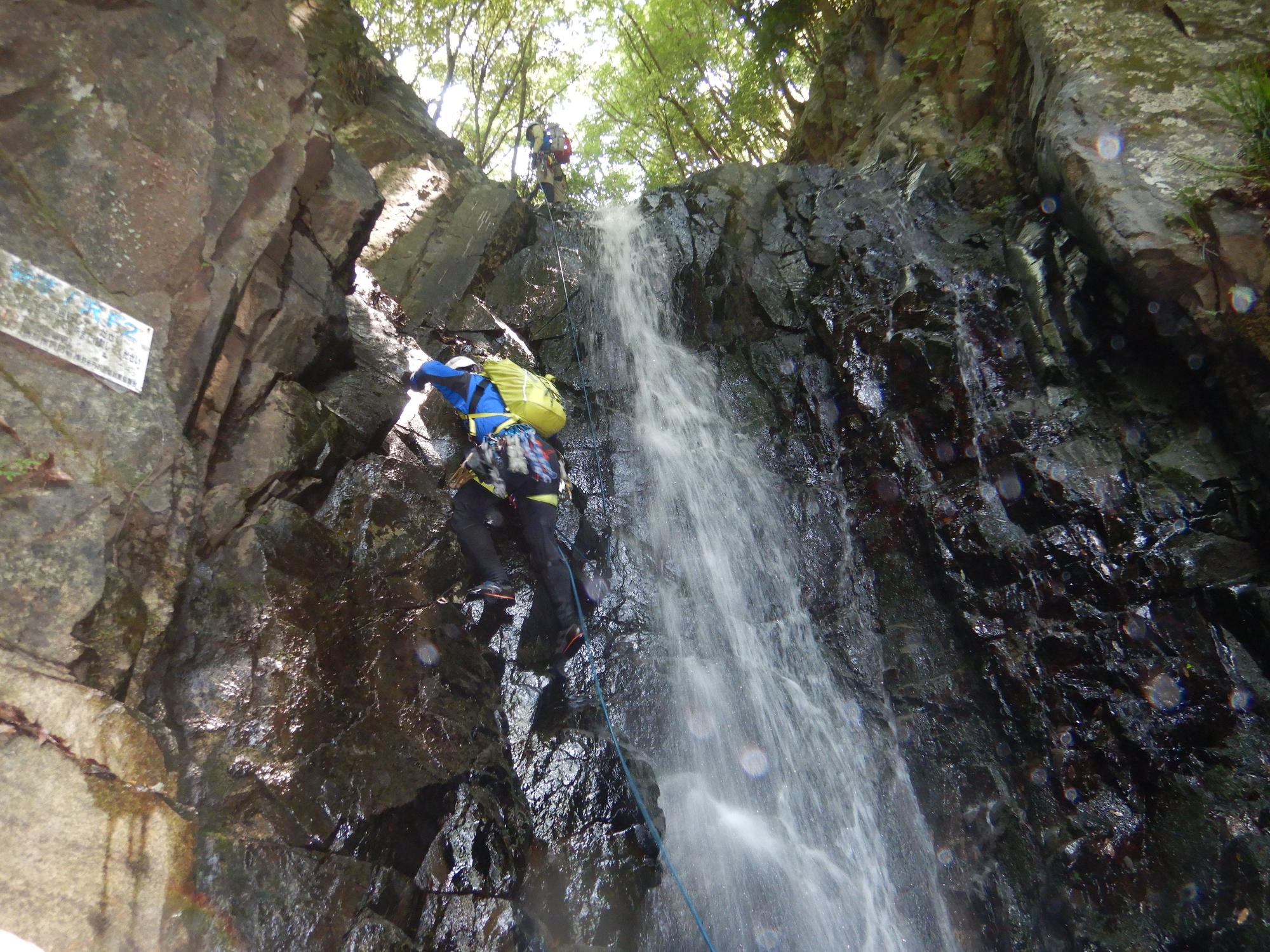 水しぶきがあがる滝の横にある急な岩壁を、青いウェアと黄色いザックを身に着けた登山者がロープを使って登り、上部にはもう一人の姿が見える沢登りの訓練の写真