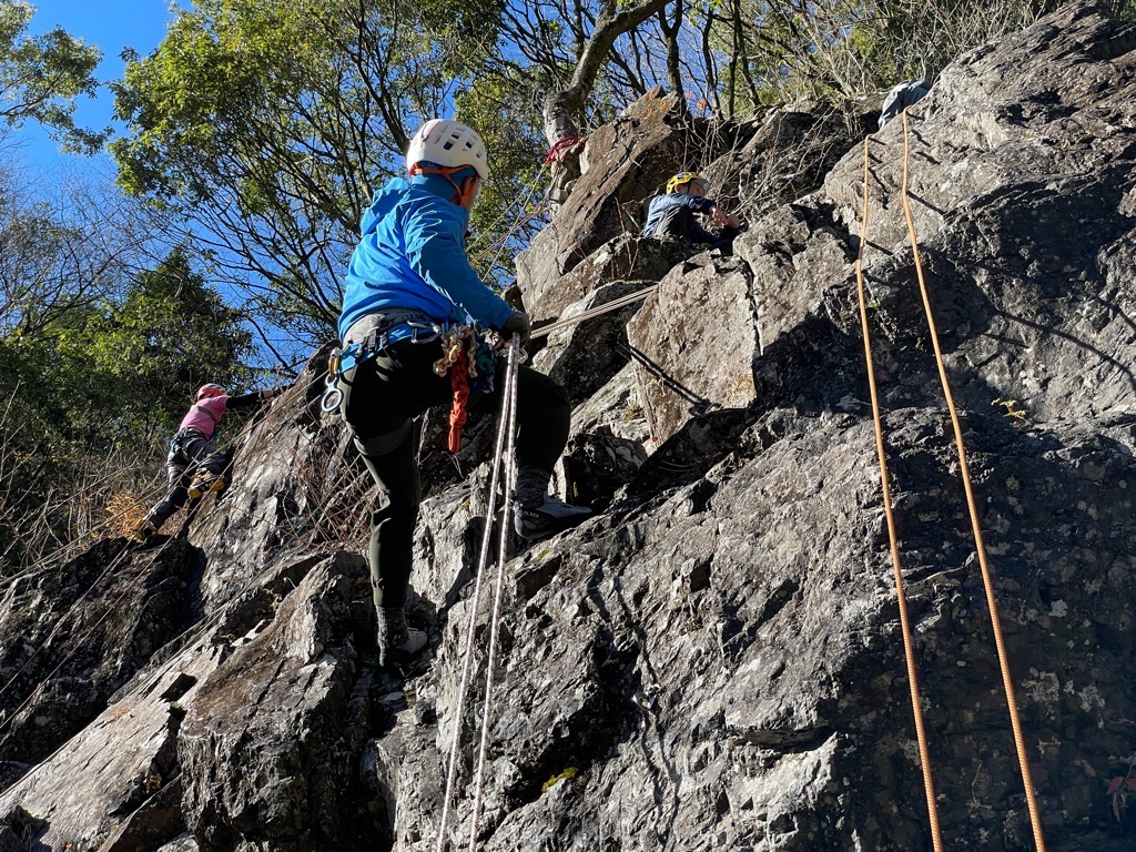 険しい岩壁でヘルメットとハーネスを装着した青いウェアの人物がロープを使ってクライミング訓練をしている写真