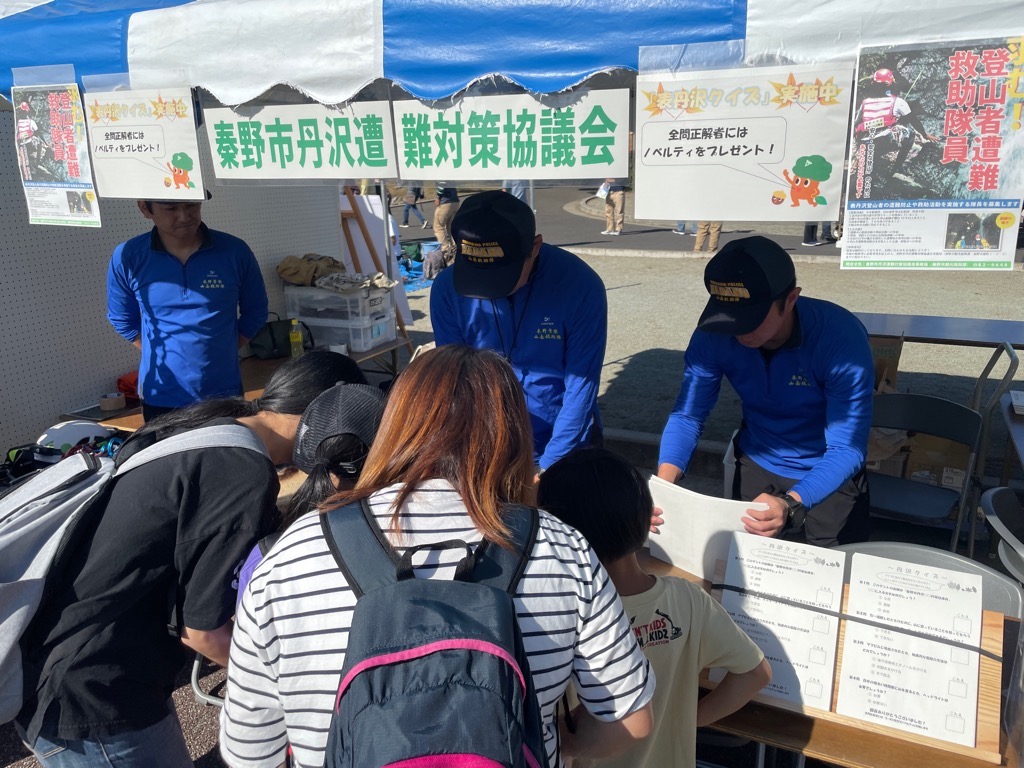 「秦野市丹沢遭難対策協議会」のテントで、青いユニフォームのスタッフが見守る中、リュックを背負った参加者たちが「全問正解者にはノベルティ」と掲示された「表丹沢クイズ」に参加しているイベントの様子の写真