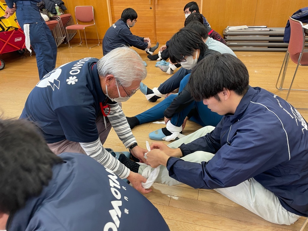 「HADANO」のロゴが入った服を着た指導員が、床に座った参加者に足首への三角巾の巻き方を指導し、周囲でもペアで練習が行われている応急手当講習の様子の写真