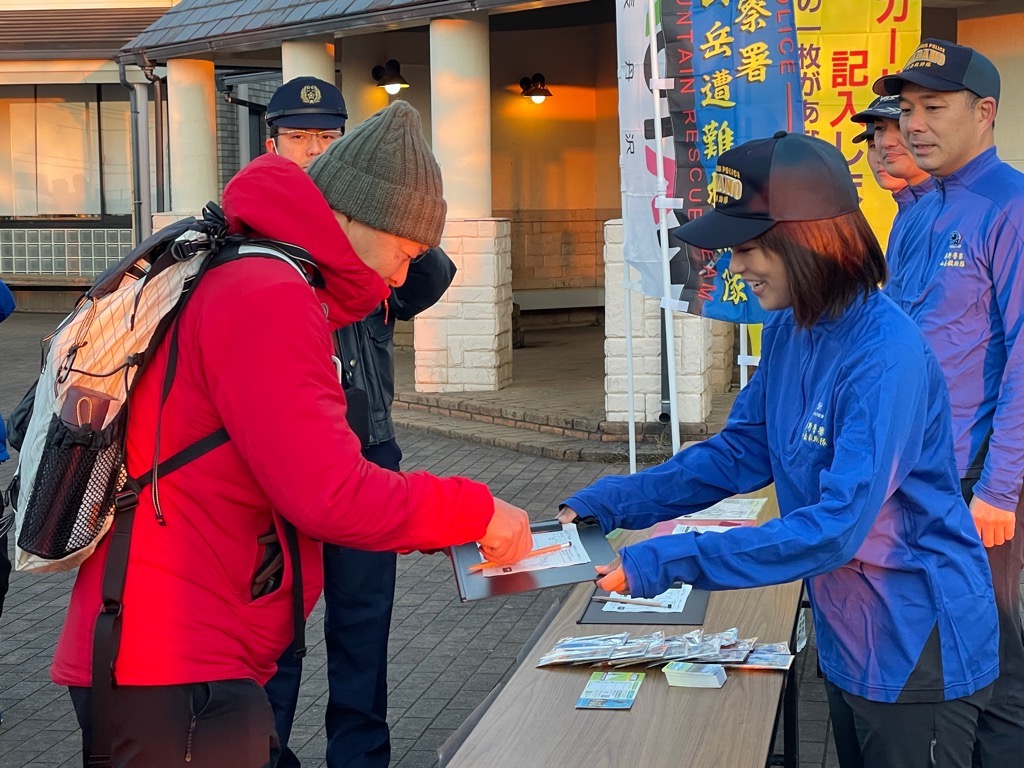 山岳救助隊などののぼり旗が立つ場所で、赤いウェアにリュック姿の登山者が、青いユニフォームを着たYouTuberのかほさんからクリップボードを受け取り、書類に記入しようとしている啓発活動の様子の写真
