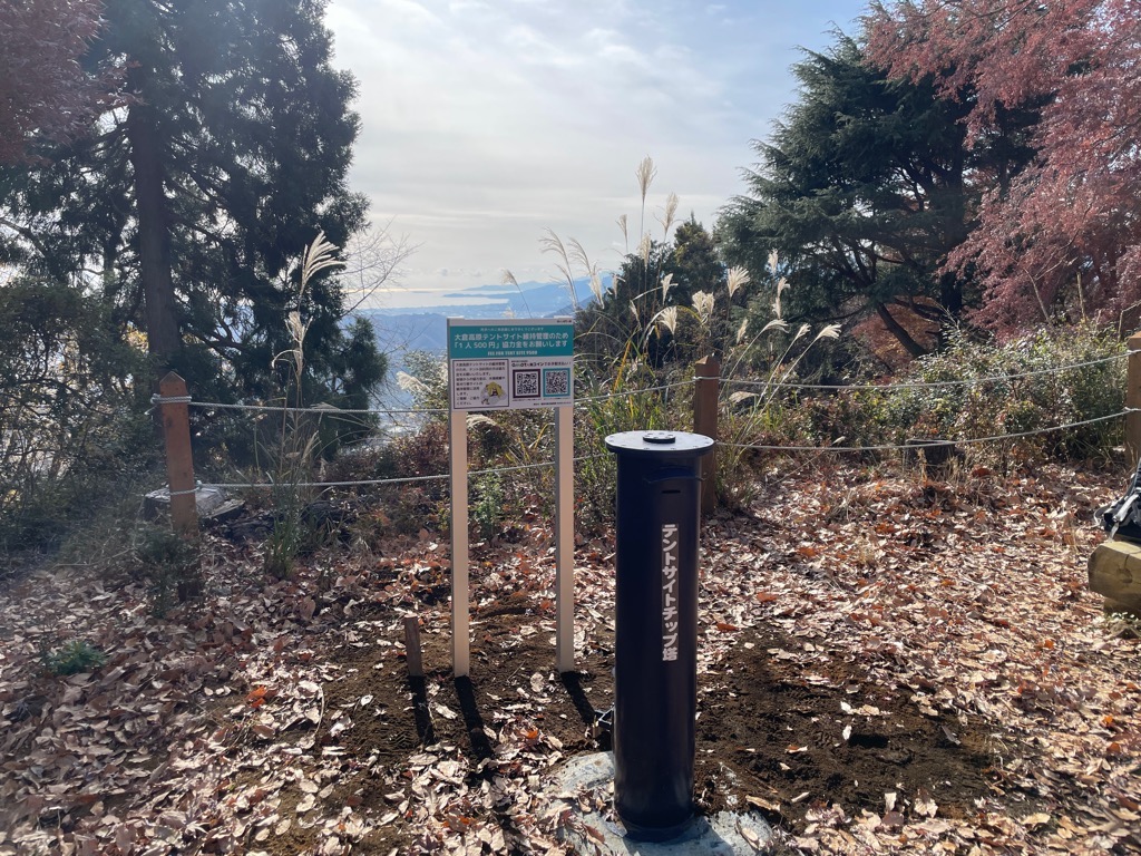 落ち葉が広がる山の上に「テントサイトチップ塔」と書かれた黒い筒と協力金を求める看板が設置されている写真