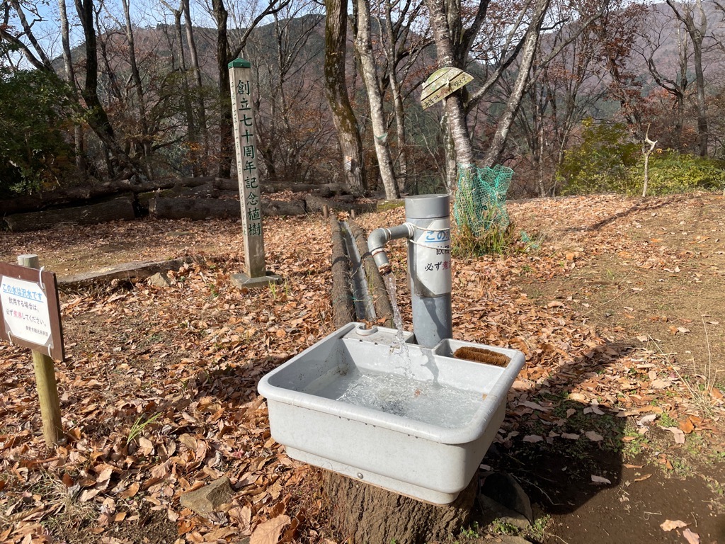落ち葉が広がる山中の広場で、「創立七十周年記念植樹」の標柱の傍らにある、「必ず煮沸」との注意書きが貼られた塩ビ管の蛇口から簡易シンクへ水が流れ落ちている写真