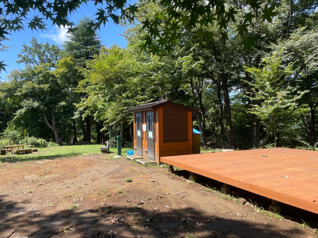 青空の下、緑豊かな木々に囲まれた広場で、手前には木製デッキが広がり、奥には茶色い外壁のトイレ棟とテーブルベンチが設置されている写真