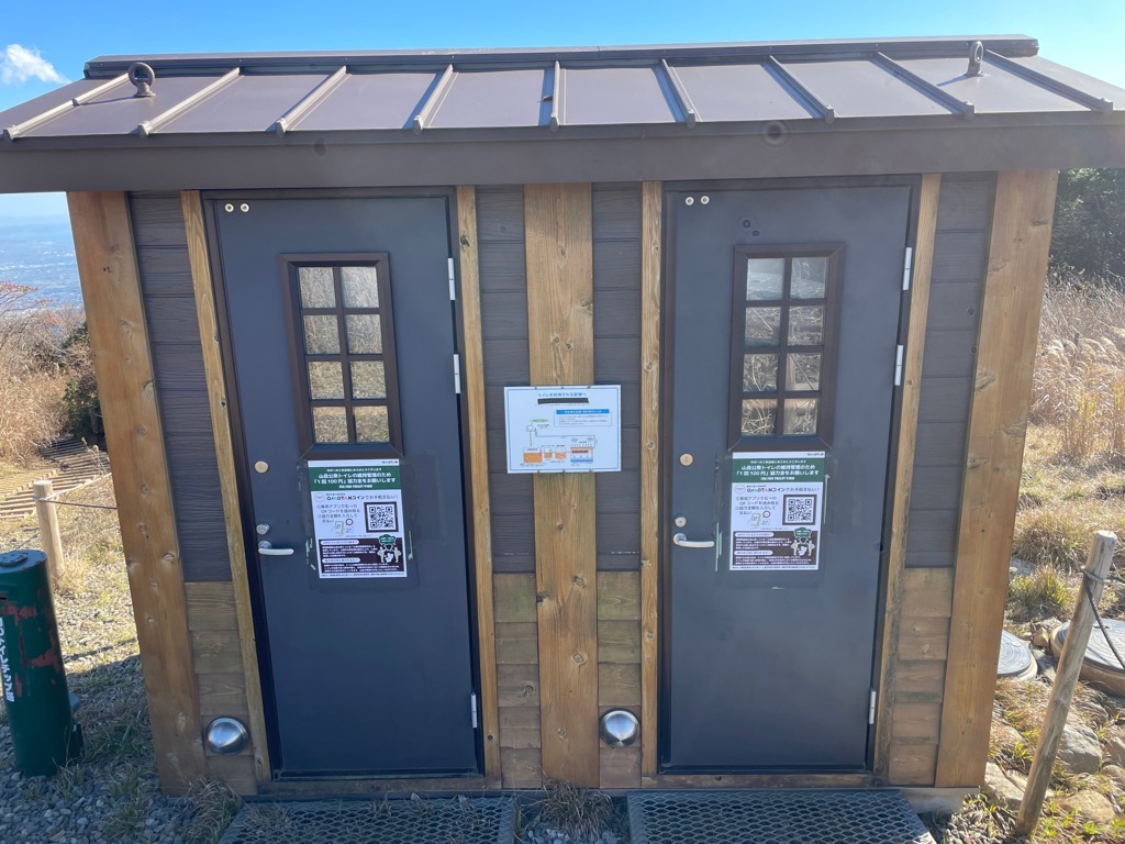 山の上に建つ木目調の公衆トイレで、2つの黒いドアには維持管理協力金を求めるQRコード付きの案内が貼られ、左端には緑色の筒が設置されている写真