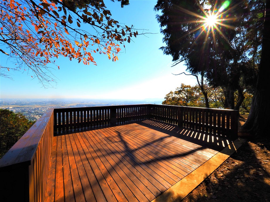 晴れ渡る青空の下、山の上にある木製展望デッキの床に柵の影が落ち、左には紅葉の枝が、右には木々越しに太陽が眩しく輝き、眼下に広大な街並みを望む風景の写真