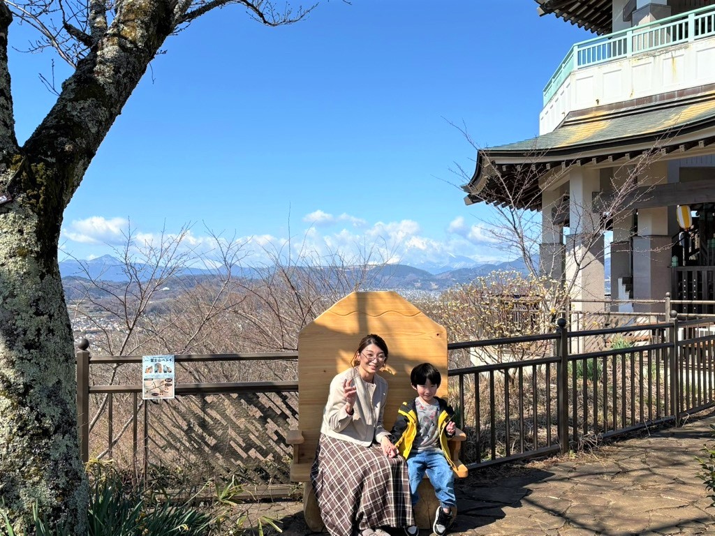 晴れ渡る青空の下、公園の展望台にある家の形をした木製ベンチに、笑顔の女性とピースサインをする男の子が座り、背景には街並みと山々が広がる写真