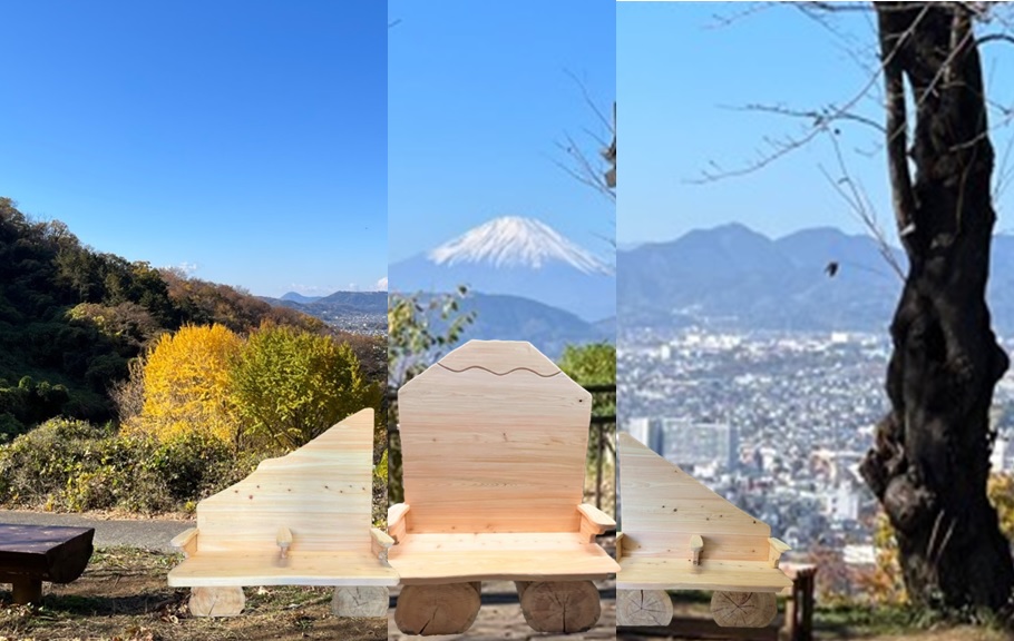 青空が広がる見晴らしの良い公園で、家の形をした背もたれが特徴的な木製ベンチが中央に置かれ、背景には左に黄葉する木々、右に市街地と山並み、そして遠くに雪を頂いた富士山が望める写真
