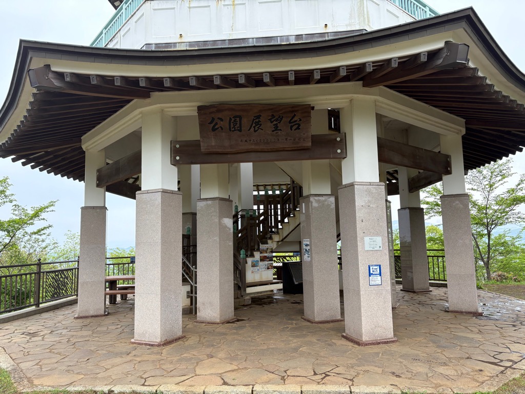 石畳の床に太い石柱が並ぶ展望台の1階部分で、奥には上階への階段が見え、中央の梁には「公園展望台」と書かれた木製の看板が掲げられている、ワイファイが設置されている権現山山頂広場の写真