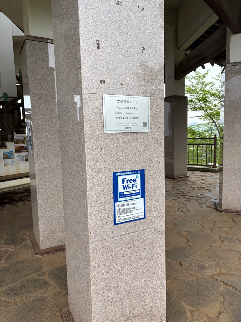 弘法山公園展望台の石畳の床に立つ御影石の太い柱に、「眺望ポイント」と記された白い案内板と、「Free Wi-Fi（ワイファイ）」と書かれた青いステッカーが並んで貼られている様子の写真