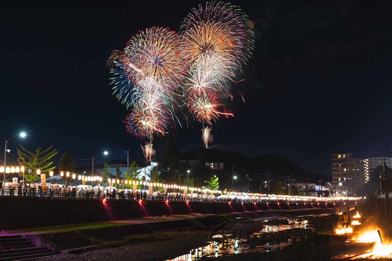 夜空に色とりどりの花火が打ち上がり、川沿いには提灯と人々でにぎわうたばこ祭の様子を写した写真
