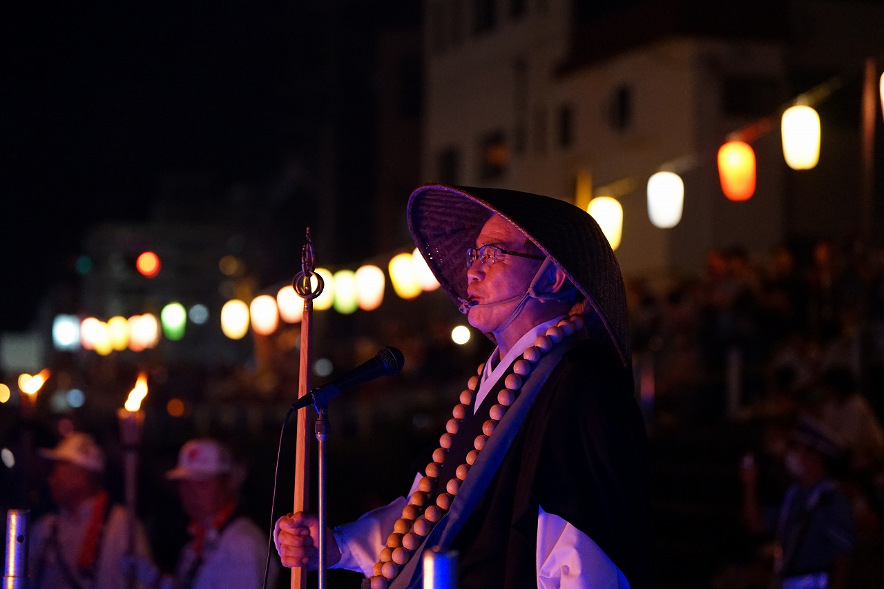夜の祭りの中、僧衣をまとい数珠をかけた僧侶がマイクの前に立っている写真