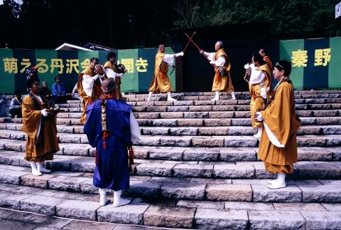 屋外の石段の舞台で、山吹色や青色の日本の伝統的な衣装をまとった僧兵たちが、棒を使った武術の演武を披露している丹沢山開きの様子写真