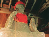 木造の建物内部に安置された赤い頭巾とよだれかけを身につけ、片手に杖を持って穏やかな表情で座っている大きな延命地蔵を下から見上げた写真