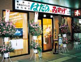 入り口上部に「はだの みやげ」と大きく書かれた看板が掲げられ、たくさんの開店祝いのスタンド花が置かれている秦野名産センター入り口付近の写真