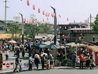 昼間の屋外広場で開催されている祭りで、大勢の人々で賑わい、テントやパラソルが立ち並び、空には赤い提灯が連なって飾られている活気ある様子の写真