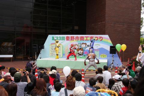 屋外イベントのステージで、赤や黄色などのカラフルなヒーローたちがアクションポーズを決めており、そのショーを子供を含む大勢の観客が柵越しに集まって見ている写真