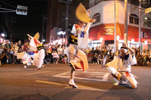 夜の市街地で、独特な形の笠や法被といった伝統衣装をまとった踊り手たちが、沿道を埋め尽くす観客の前で躍動的に踊っている写真